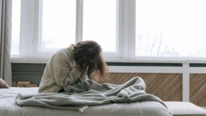 Person sitting in bed in morning light, head lowered, conveying exhaustion rather than sadness